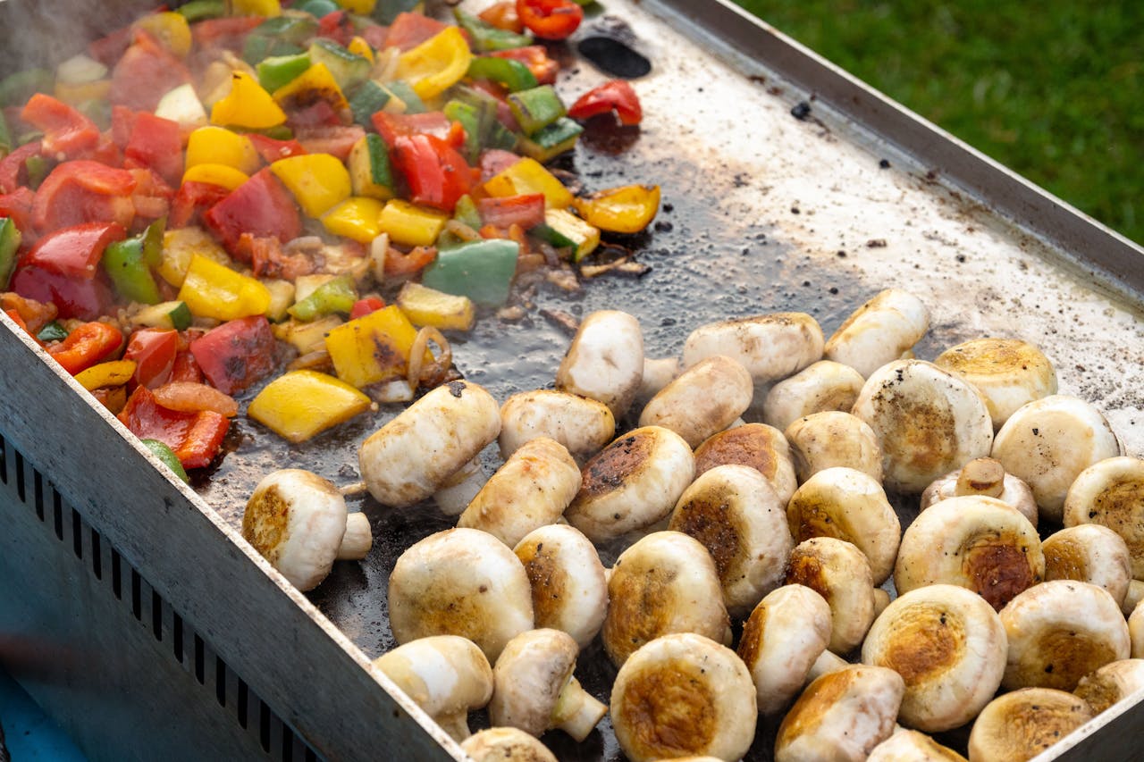 Colorful grilled vegetables and mushrooms sizzling on a barbecue grill plate outdoors.