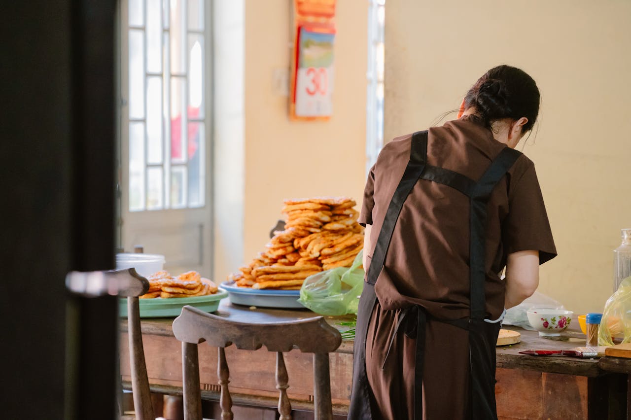 A woman in a brown apron prepares food beside a stack of pancakes in a sunlit kitchen.